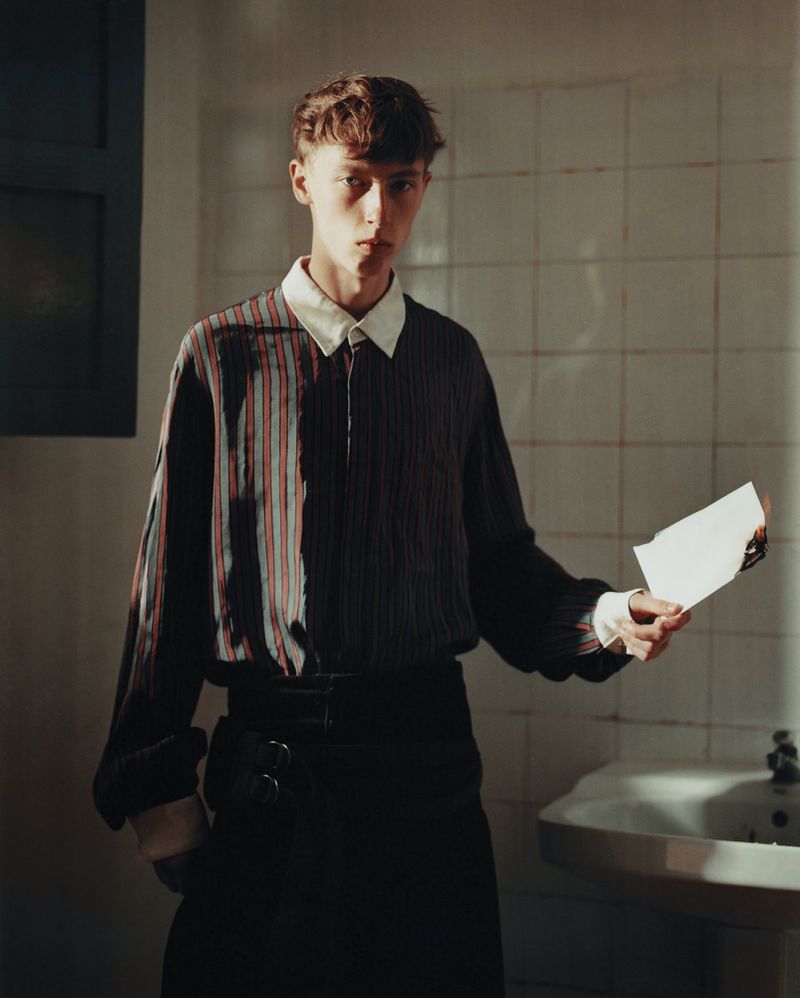 Young man in a striped collared shirt holding a sheet of white paper in a tiled bathroom beside a sink.