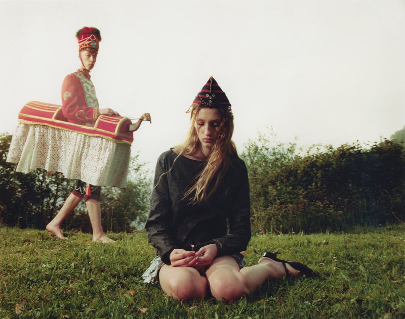 Woman in a pointed hat kneeling in a meadow with a figure wearing a theatrical hobby horse costume in the background.