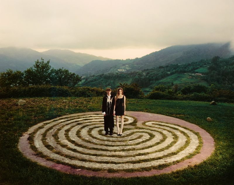 Two fashion models in black clothing stand at the center of a circular stone labyrinth on a grassy hill overlooking misty mountains.