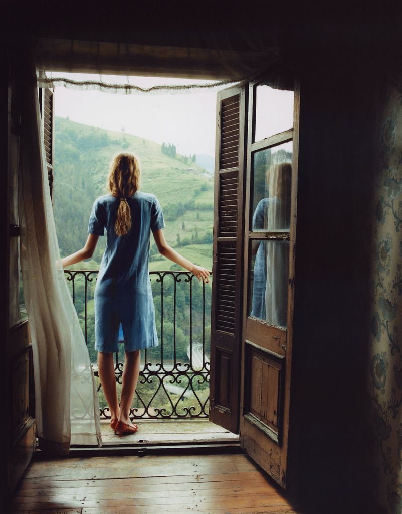 A woman in a blue short-sleeve dress stands on a wooden balcony overlooking green mountains through open shuttered doors.