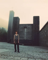 Model wearing a grey tank top and dark trousers standing in a cobblestone courtyard before a brutalist stone church in misty weather.