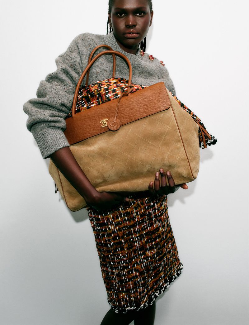 Model Nyibol Dok Jok holding a large camel suede and brown leather tote bag wearing a tweed skirt against a white studio background.