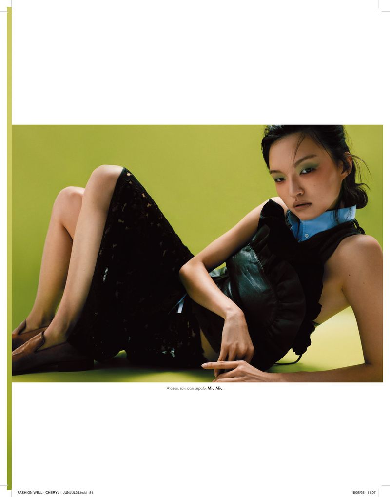 Model reclining on a vibrant green studio backdrop wearing a black ruffled top, dark midi skirt, and brown loafers for Elle Indonesia.