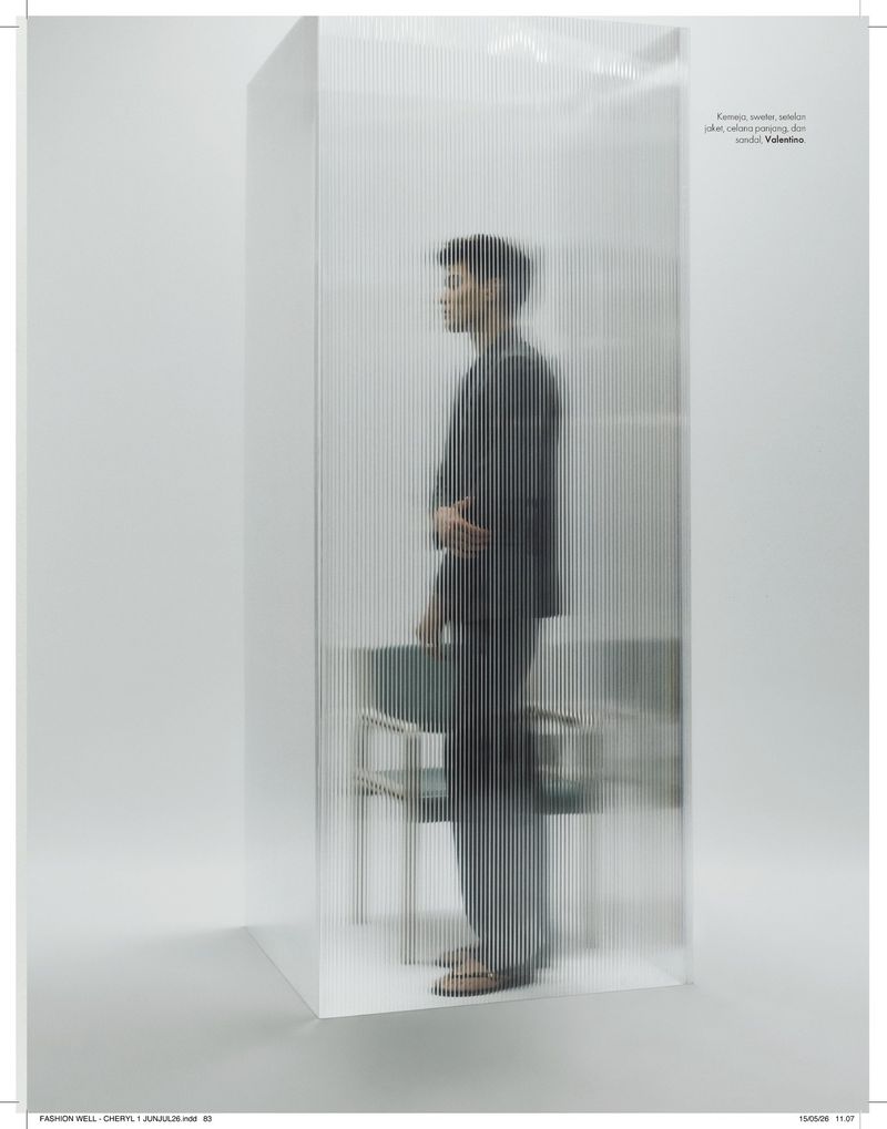 Model standing behind a translucent ribbed glass screen wearing a dark suit in a minimal studio setting.