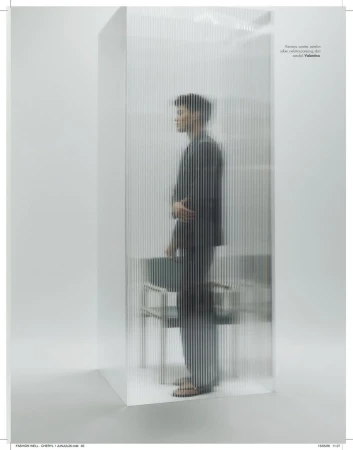 Model standing behind a translucent ribbed glass screen wearing a dark suit in a minimal studio setting.