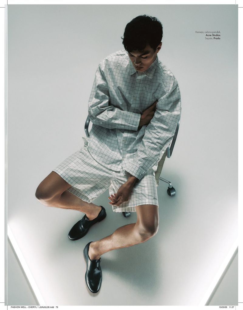 High-angle shot of a man seated in an office chair wearing a matching grid-pattern shirt and shorts with black leather dress shoes.