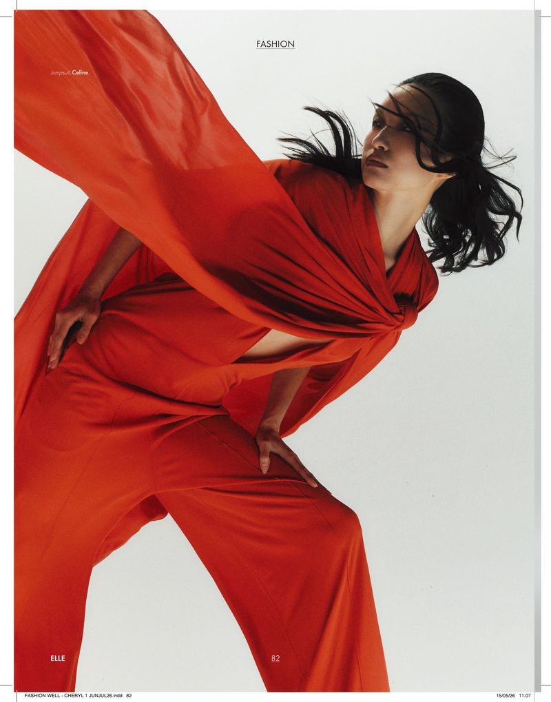 Model posing dynamically in a flowing draped orange jumpsuit with windswept hair against a white background.