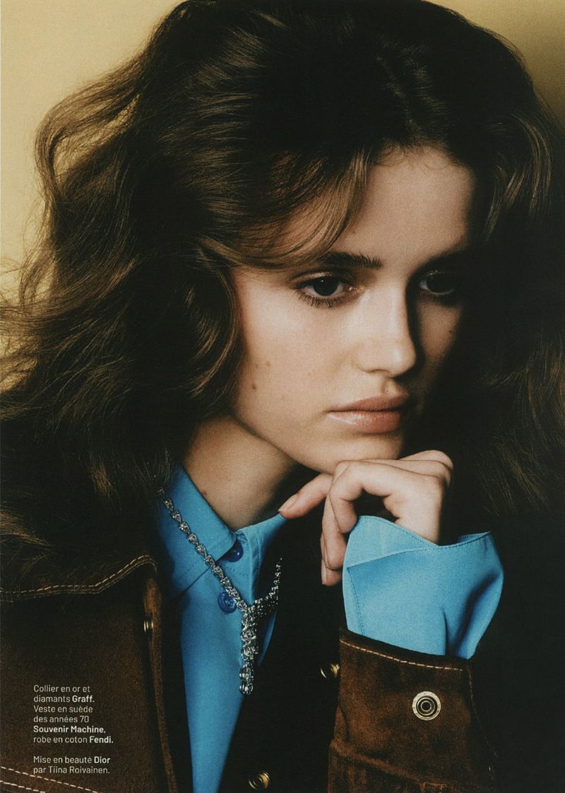 Floor Vrins resting her chin on her hand wearing a brown suede jacket, blue shirt, and diamond necklace against a warm backdrop.