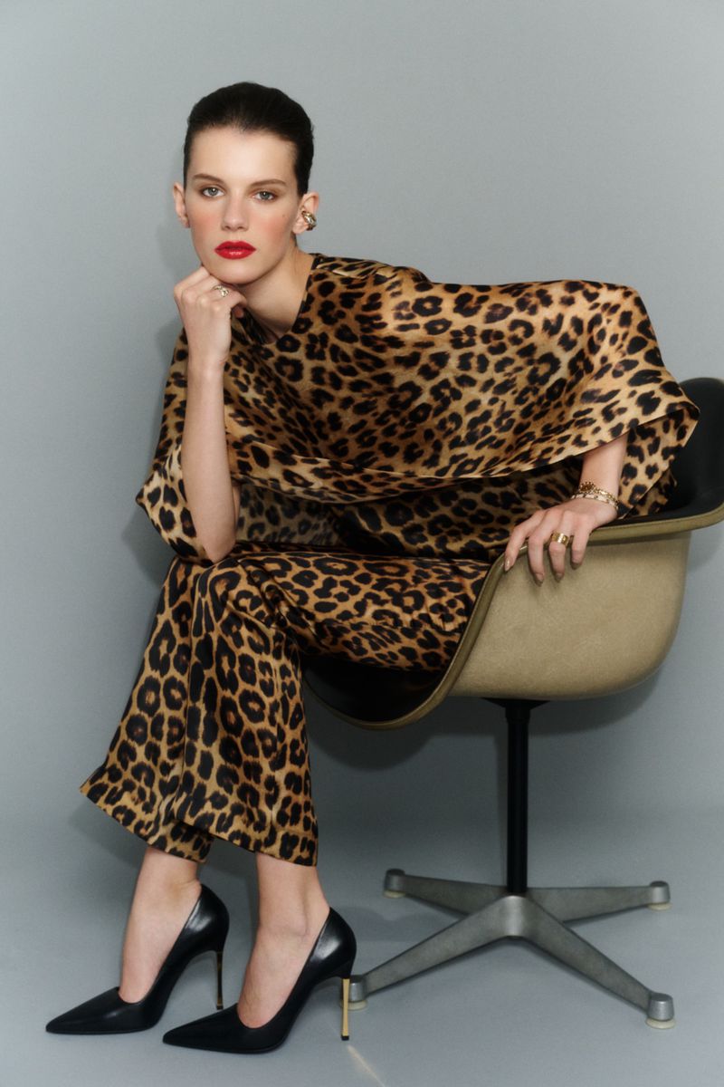 Model wearing a leopard print outfit seated in an office chair against a grey studio backdrop