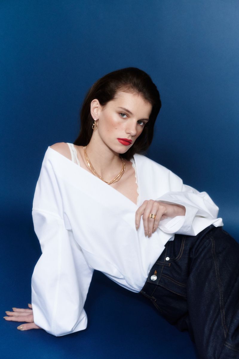 Paula Engbert reclining in an off-the-shoulder white blouse, dark jeans, and gold jewelry against a deep blue studio backdrop.