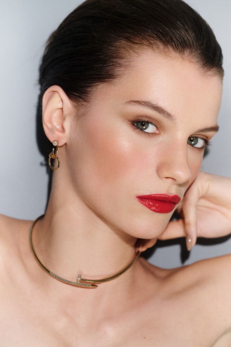 Close-up portrait of model Paula Engbert wearing bold red lipstick, gold drop earrings, and a gold nail-inspired choker necklace.