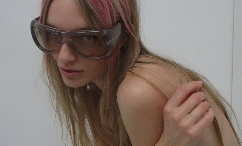 Model Maya Stepper wearing oversized translucent tinted shield sunglasses against a plain white studio backdrop.