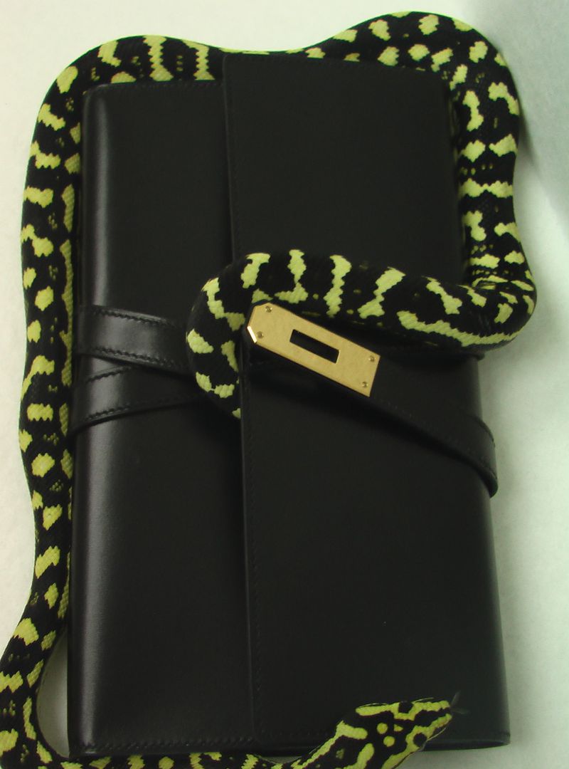 Black leather clutch with gold turn-lock hardware wrapped by a yellow and black patterned snake on a pale neutral backdrop.