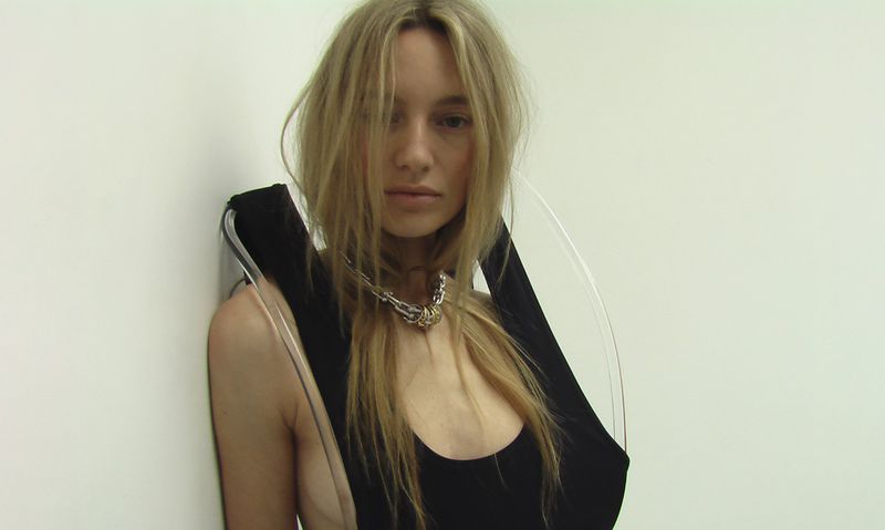Maya Stepper wearing a black sleeveless garment with curved transparent acrylic accents against an off-white background.