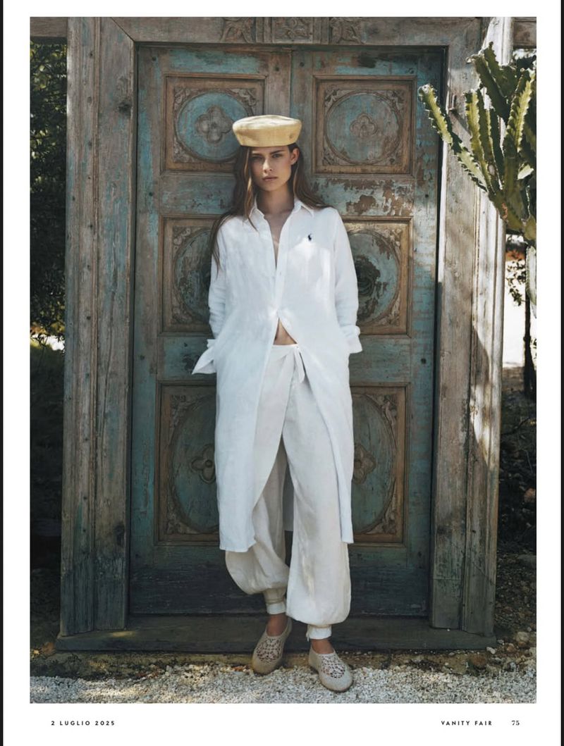 A woman in a white linen outfit and straw hat stands in front of an ornate teal wooden door, with a sunlit cactus to her right.