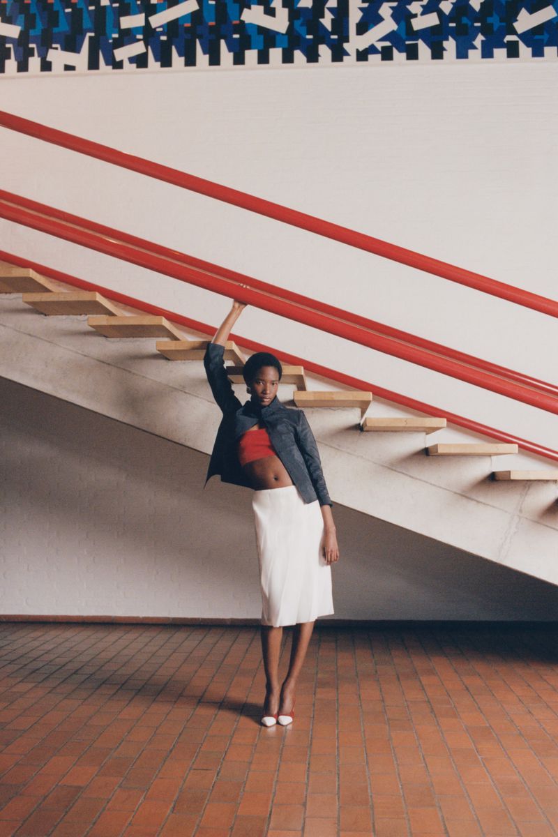 Model Zayna Cisse wearing a white midi skirt and jacket posing beneath a red-railed staircase for Eurowoman.