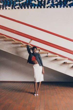 Model Zayna Cisse wearing a white midi skirt and jacket posing beneath a red-railed staircase for Eurowoman.