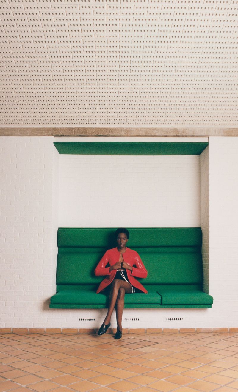 Model Zayna Cisse sitting on a green built-in bench wearing a red jacket and black shoes against a white alcove wall.