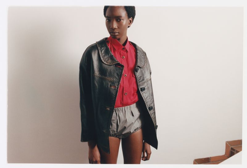 Model Zayna Cisse in an oversized black leather jacket, red collared shirt, and grey shorts posing against a light neutral wall.
