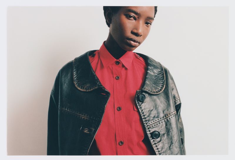 Model Zayna Cisse wearing a bright red collared shirt under a dark distressed leather jacket against a white studio background.
