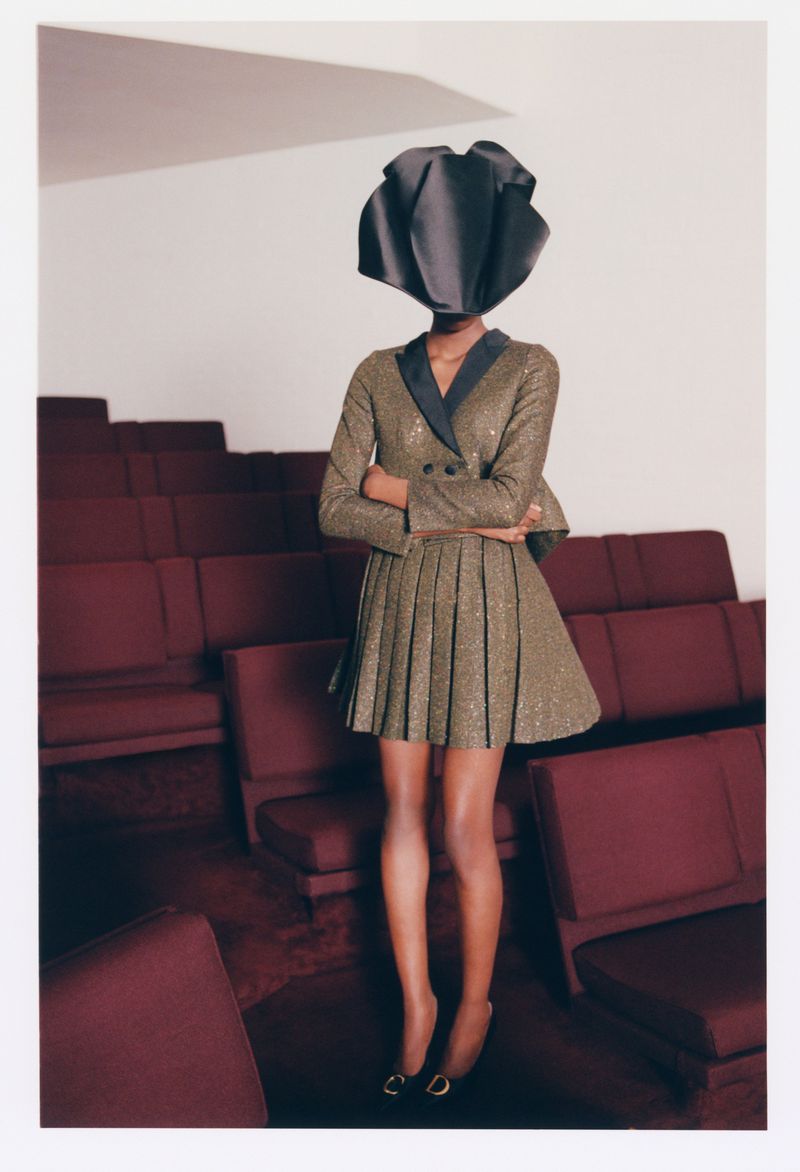 Model with a sculptural black headpiece wearing a patterned blazer and pleated skirt in an auditorium with red seats.