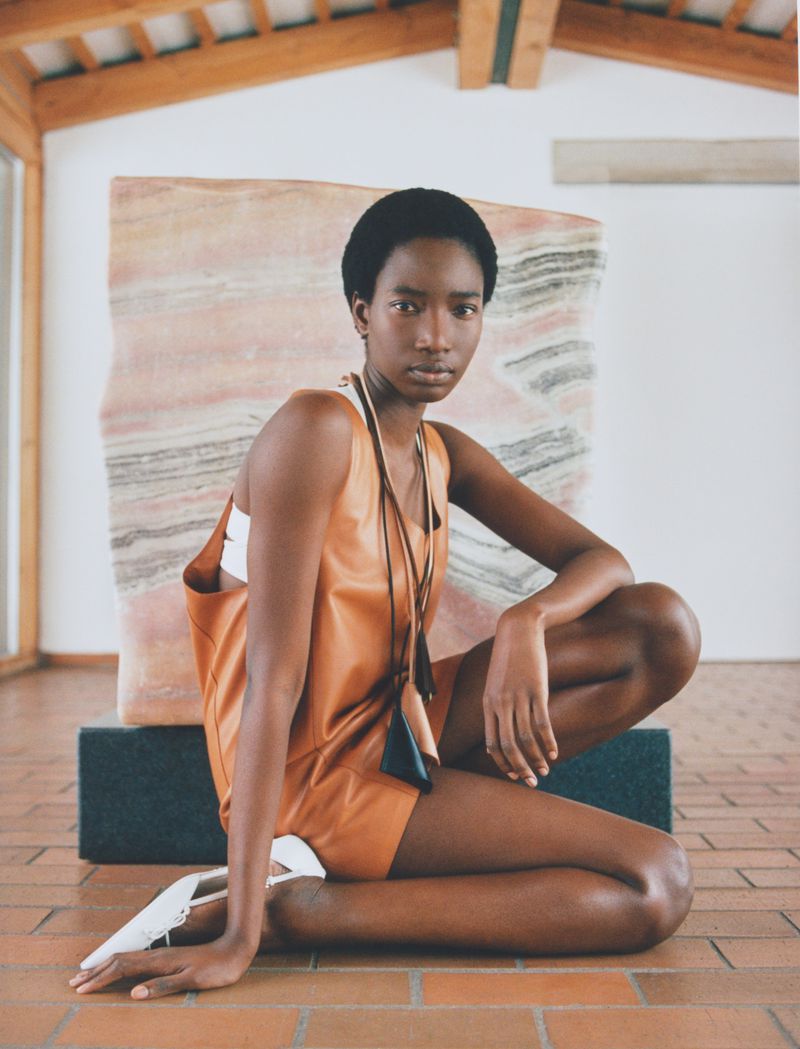 Model Zayna Cisse crouching in a tan leather dress and white shoes before a stone slab on a terracotta tile floor.