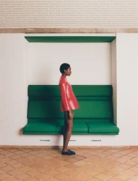 Model Zayna Cisse standing in profile wearing a structured red short dress in front of a green recessed bench on terracotta tiles.