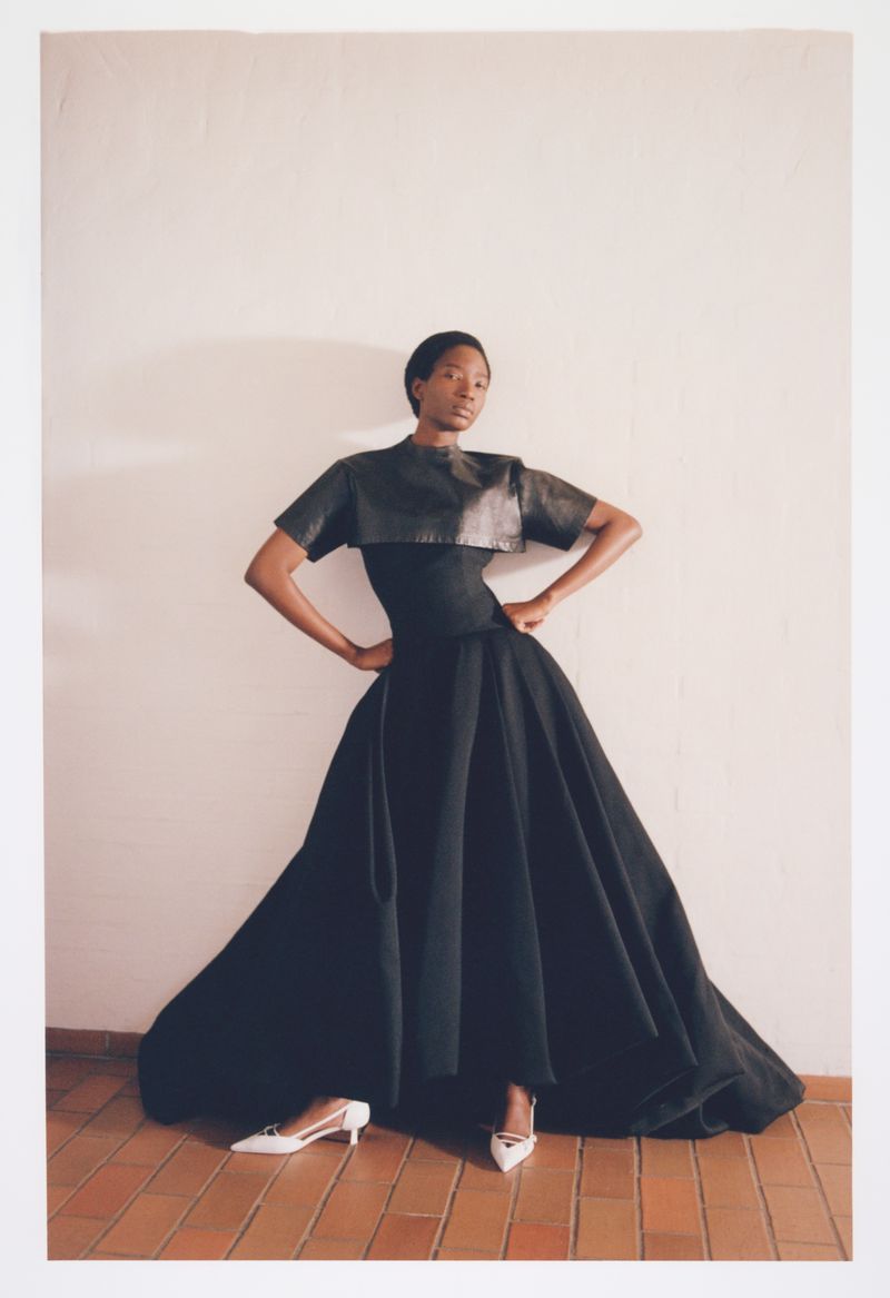 Model Zayna Cisse poses in a voluminous black gown with a cropped leather top and white slingback heels against an off-white wall.