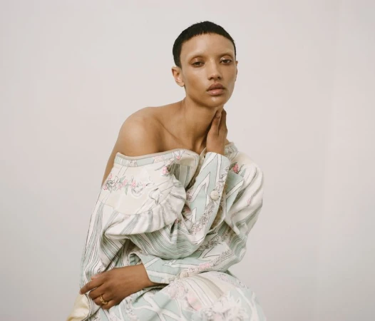 Model Georgia Palmer posing in a draped off-shoulder pastel patterned coat against an off-white seamless backdrop.