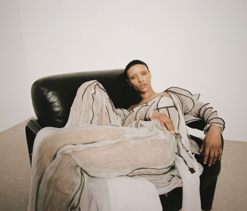 Model Georgia Palmer reclines in a black leather armchair wearing a draped grey gown against a white backdrop.