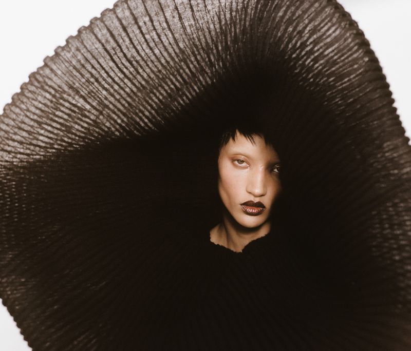 Model Georgia Palmer wearing an oversized circular pleated black sculptural piece around her face against a bright background.