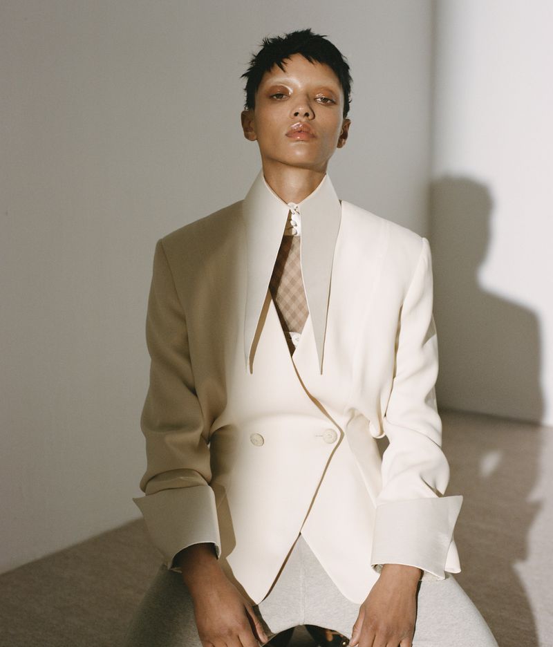 Model Georgia Palmer seated wearing an off-white structured blazer with exaggerated pointed lapels in a studio setting.