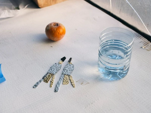 Still life with artistic drop earrings, an orange fruit, and a clear plastic cup of water on a light tabletop in natural sunlight.