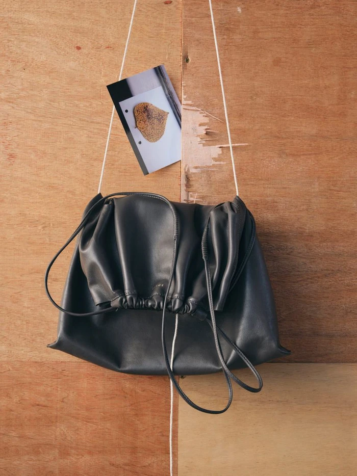 Black leather drawstring pouch bag suspended by white strings against a wood panel backdrop with a small printed photograph above.