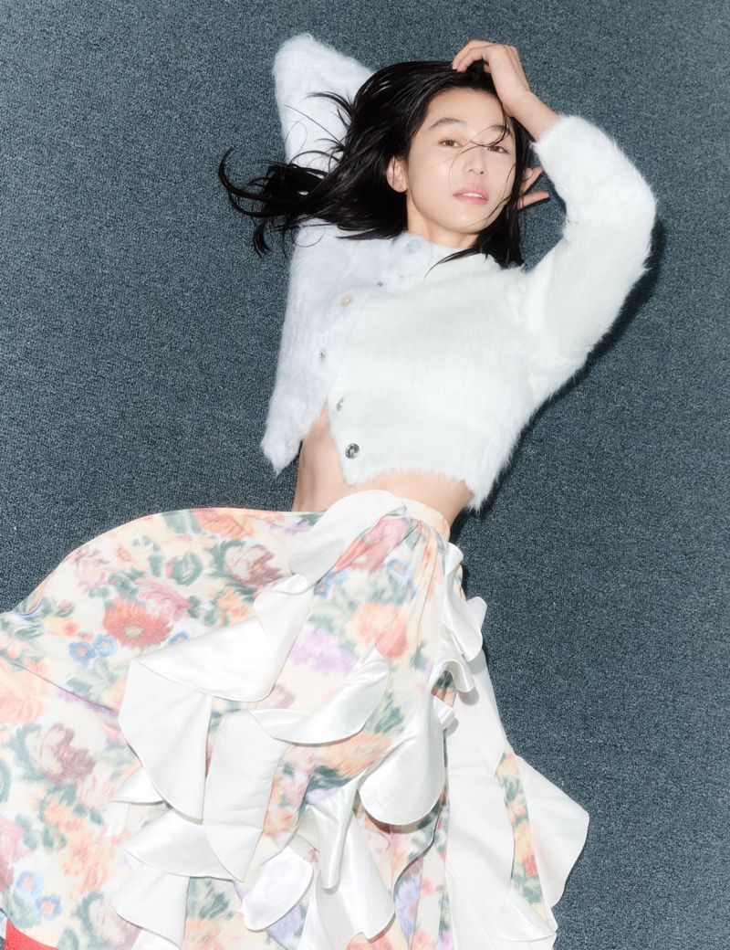 Gianna Jun lying on a textured grey surface wearing a white cropped fuzzy sweater and floral ruffled skirt.