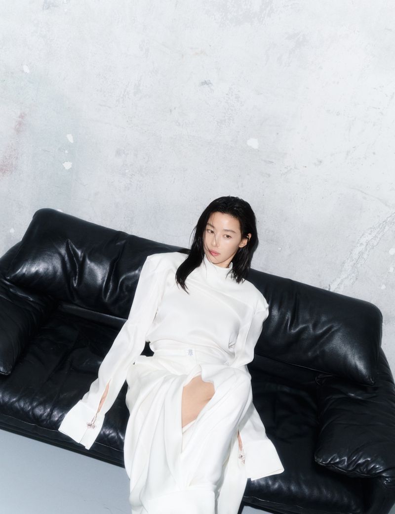 Jun Ji-hyun wearing an all-white ensemble seated on a black leather sofa in front of a textured wall