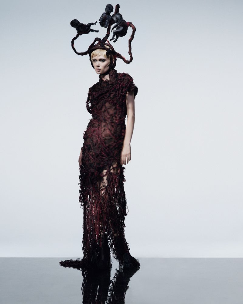 Model Puck Loomans wearing a textured dark red shredded dress and an avant-garde sculptural headpiece in a minimalist studio setting.
