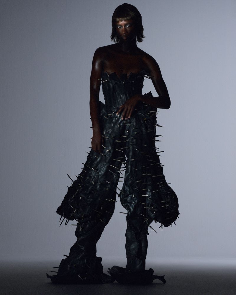 Model Puck Loomans posing in a black spiked sculptural outfit against a dark studio gradient backdrop for Glamcult Magazine.