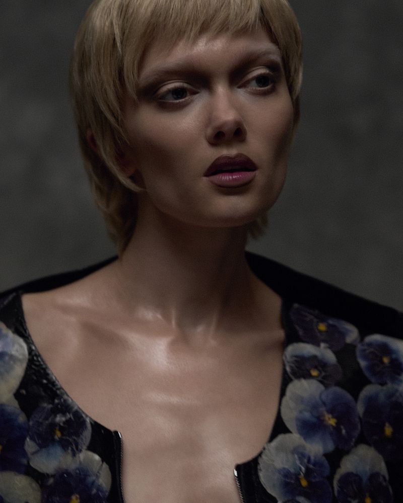 Model Puck Loomans with short blonde hair wearing a floral printed black jacket against a dark textured backdrop.