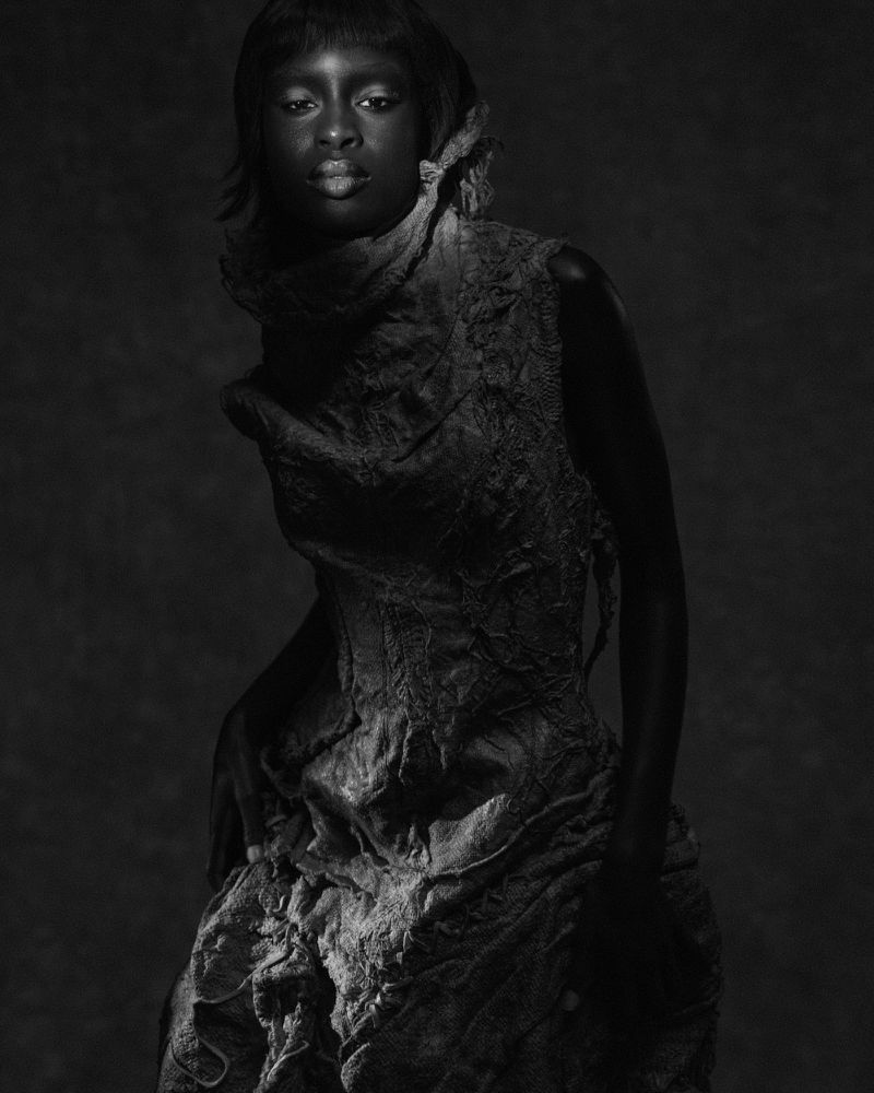 Black and white portrait of a Black woman posing in a heavily textured, distressed sleeveless high-neck garment against a dark backdrop.