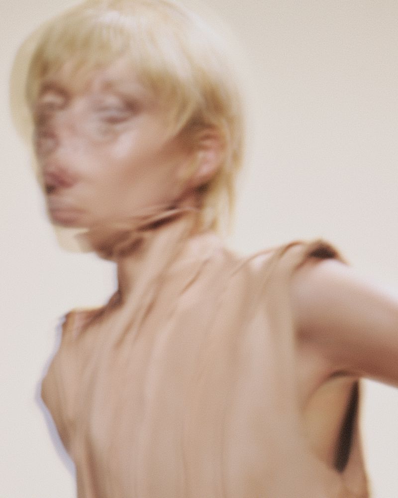 Motion-blurred portrait of blonde model Puck Loomans wearing a draped beige top against a warm studio background.