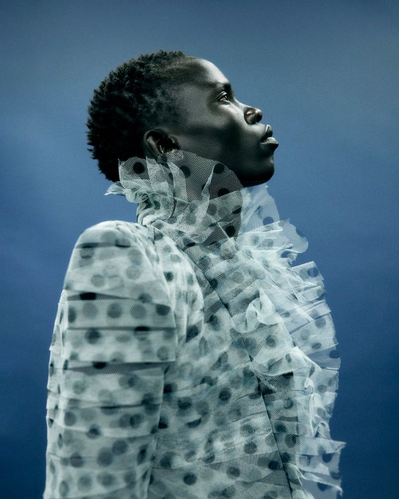 Profile portrait of model Christopher Akech wearing a pleated polka dot ruffled jacket against a blue studio backdrop.