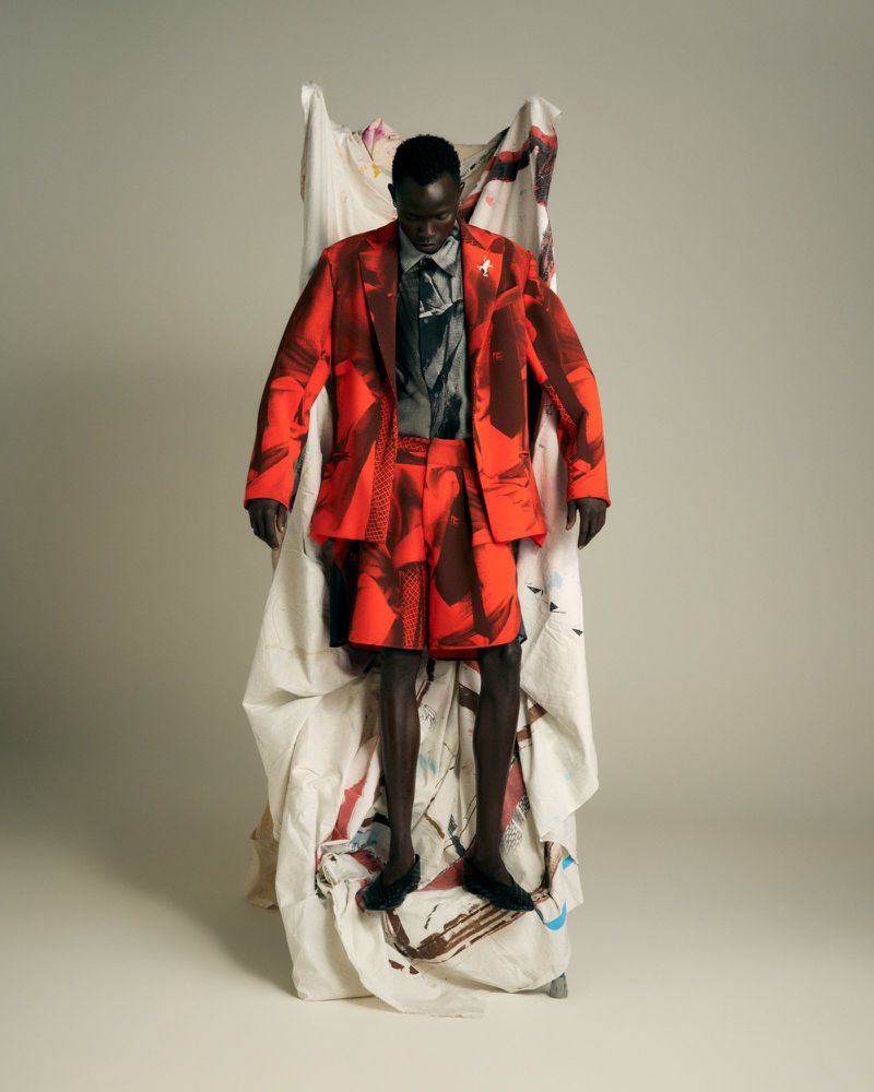 Model wearing a red patterned blazer and matching shorts standing against a draped painted canvas in a studio.