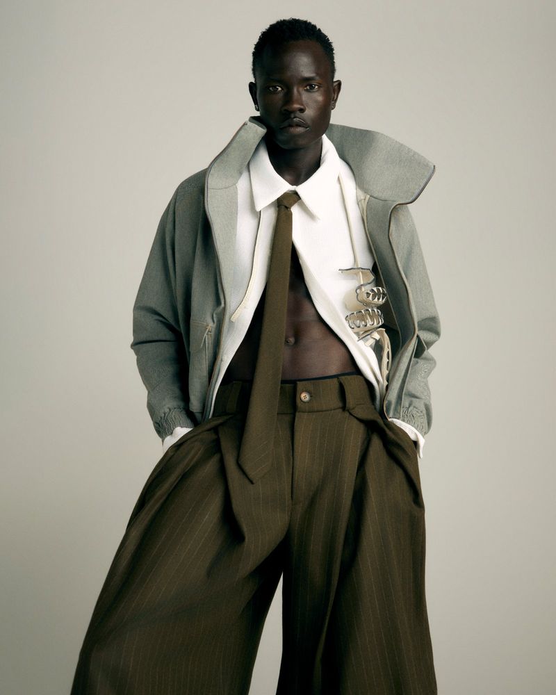 Christopher Akech wearing an open white shirt, olive tie, high-collar jacket, and wide-leg trousers against a neutral backdrop.