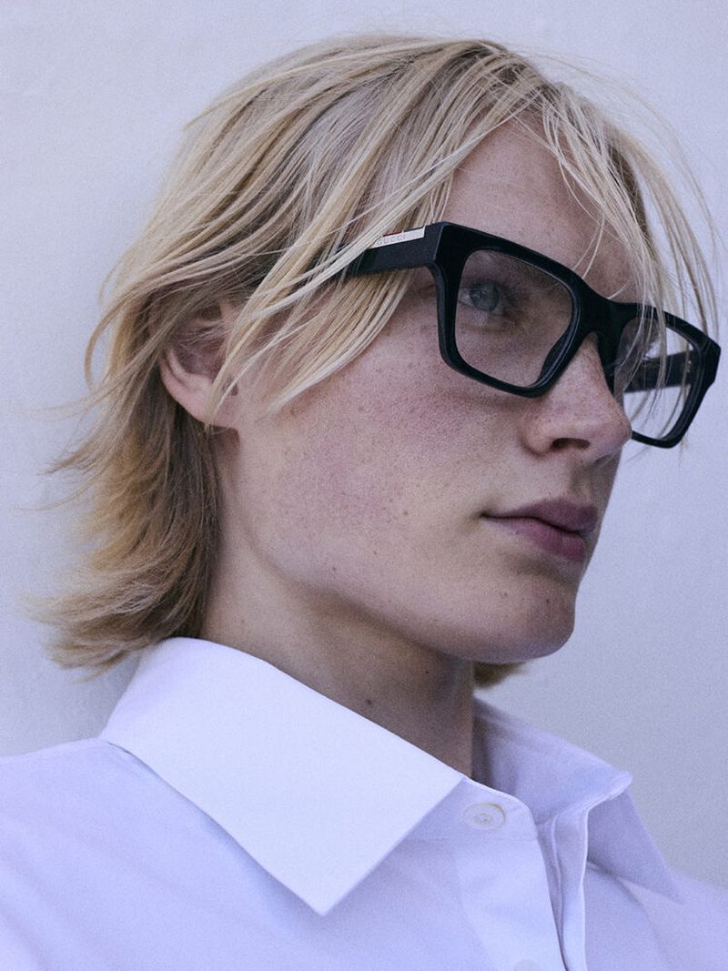Portrait of a young adult with blond hair wearing square black-rimmed glasses and a white collared shirt, set against a white backdrop.