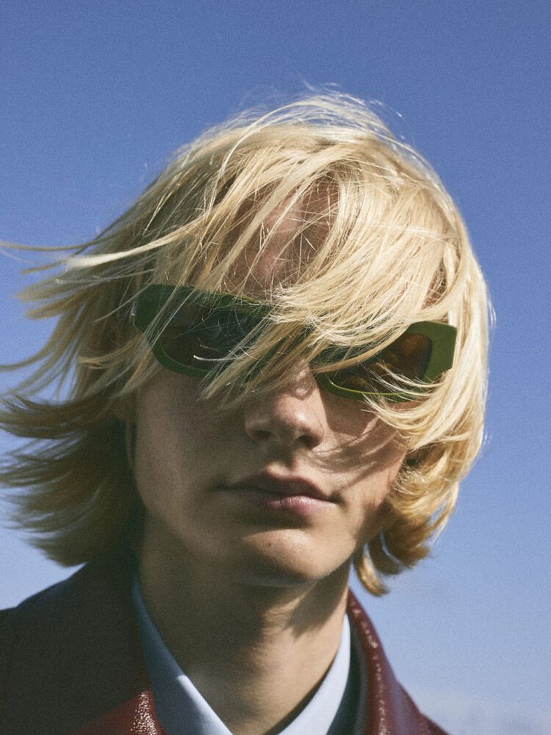 Model Leander Straub with windswept blonde hair wearing green sunglasses against a clear blue sky.