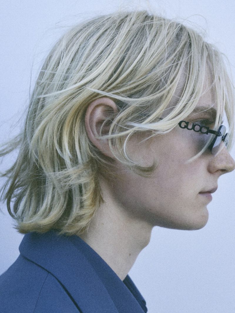Close-up shot of a person with blond hair, wearing a blue blazer and sunglasses with the Gucci logo on the frame. Side profile.