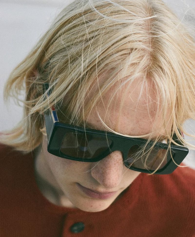 Close-up of a young adult with blonde hair and dark sunglasses, wearing a brown cardigan against a light background, focusing on fashion.