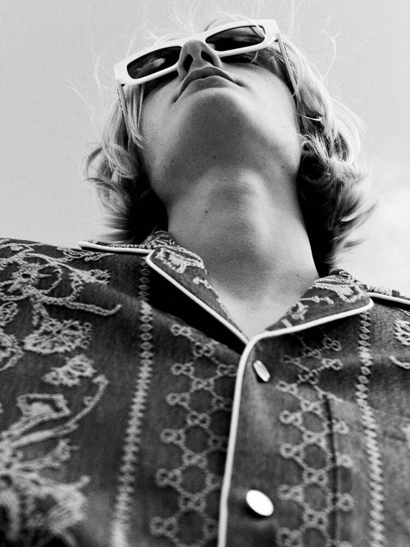 Monochrome, low-angle portrait of a fair-skinned person with blond hair wearing sunglasses and a patterned button-down shirt outdoors.