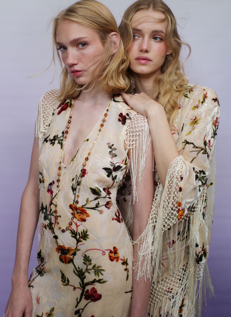 Two female models wearing floral printed dresses with fringe details posing against a neutral gradient background.